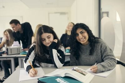 Studenter i klassrummet, en elev ler mot kameran medan en annan studerar.