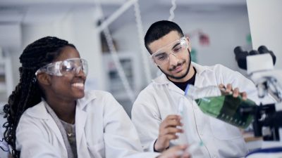 Två elever med vita rockar och skyddsglasögon laborerar med grön vätska