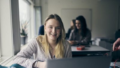Student med laptop leende i klassrum.